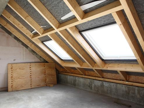 Blick in einen leeren Dachraum mit Holzrahmen, Fenster und verputzten Wänden, eine Holzplatte liegt am Boden.